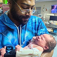 Emmett joined the competition — help win amazing prizes! man, baby, newborn, hospital, medical_equipment, glasses, blue_hoodie, facial_hair, holding, care, person, indoor, closeup, tubes, monitor, bedroom, crying, skin, arm, expression