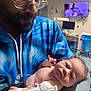 man, baby, hospital, medical_brace, iv_line, glasses, blue_hoodie, newborn, patient, care, father, child, indoor, medical_equipment, monitor, tv_screen, bedside, tender_moment, portrait, healthcare