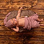 baby, infant, pacifier, beads, hat, pink_cloth, cushion, wooden_floor, lying_down, raised_arm, skin, face, eyes, hand, foot, texture, indoor, cute, soft, resting
