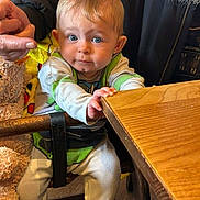 Logan is registered to the contest to win money with this photo: baby, child, blue_eyes, wooden_table, high_chair, hand, teddy_bear, blanket, clothing, person, indoor, cute, support, sitting, furniture, young_child, curious, face, expression, holding