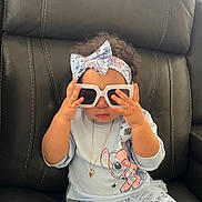 Nathasha Marie joined the competition — help win amazing prizes! toddler, child, sunglasses, headband, cartoon_clothing, black_leather_chair, sitting, indoor, cute, playing, hand, face, jewelry, necklace, curly_hair, toy, pants, shirt, window_blinds, light