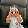 toddler, child, sunglasses, headband, cartoon_clothing, black_leather_chair, sitting, indoor, cute, playing, hand, face, jewelry, necklace, curly_hair, toy, pants, shirt, window_blinds, light