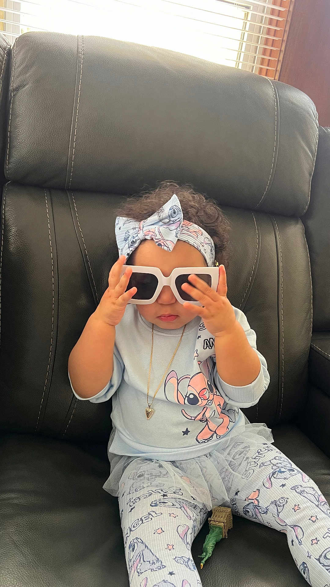 Nathasha Marie joined the competition — help win amazing prizes! toddler, child, sunglasses, headband, cartoon_clothing, black_leather_chair, sitting, indoor, cute, playing, hand, face, jewelry, necklace, curly_hair, toy, pants, shirt, window_blinds, light