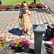 Deanna is registered to the contest to win money with this photo: child, girl, flower_bed, pavement, backpack, dress, sandals, shadow, greenery, park, sunny, outdoor, nature, plants, road, people, stone_border, pink_flowers, white_flowers, grass