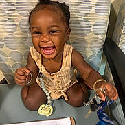 Myla is registered to the contest to win money with this photo: toddler, child, smiling, happy, indoor, chair, keys, lanyard, pacifier, paper, document, clothing, bracelet, hair, face, person, sitting, table, pattern, accessory
