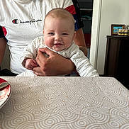 Lysaé participe au concours pour gagner de l'argent avec cette photo : baby, adult, smile, table, tablecloth, hand, clothing, indoor, furniture, person, face, portrait, pattern, beard, sitting, white_shirt, holding, cheerful, casual, home