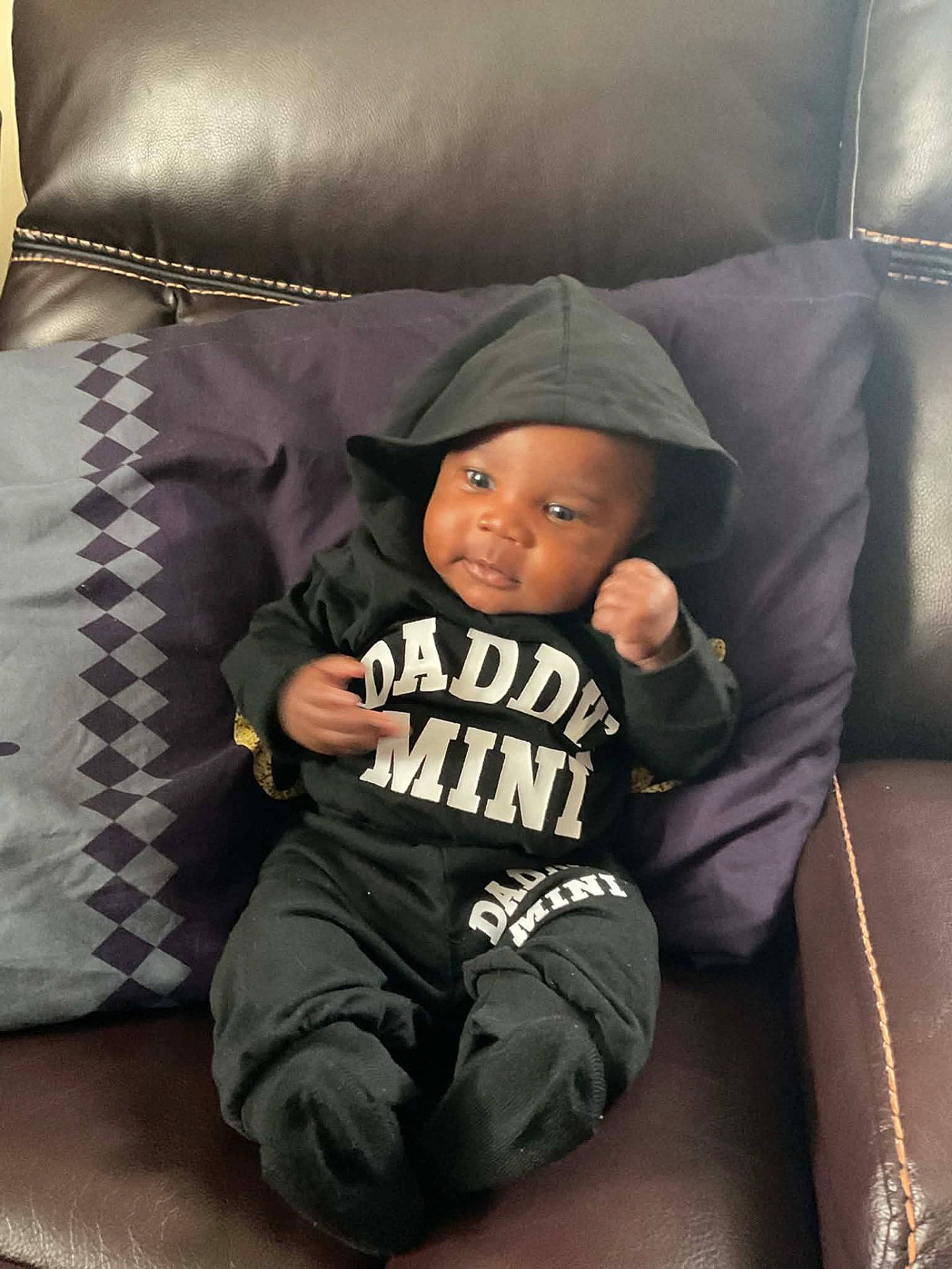 Jermany is registered to the contest to win money with this photo: baby, black_clothing, casual, child, clothing, comfortable, couch, cute, fist, hoodie, indoors, infant, leather, person, pillow, portrait, purple_pillow, sitting, smiling, young_child