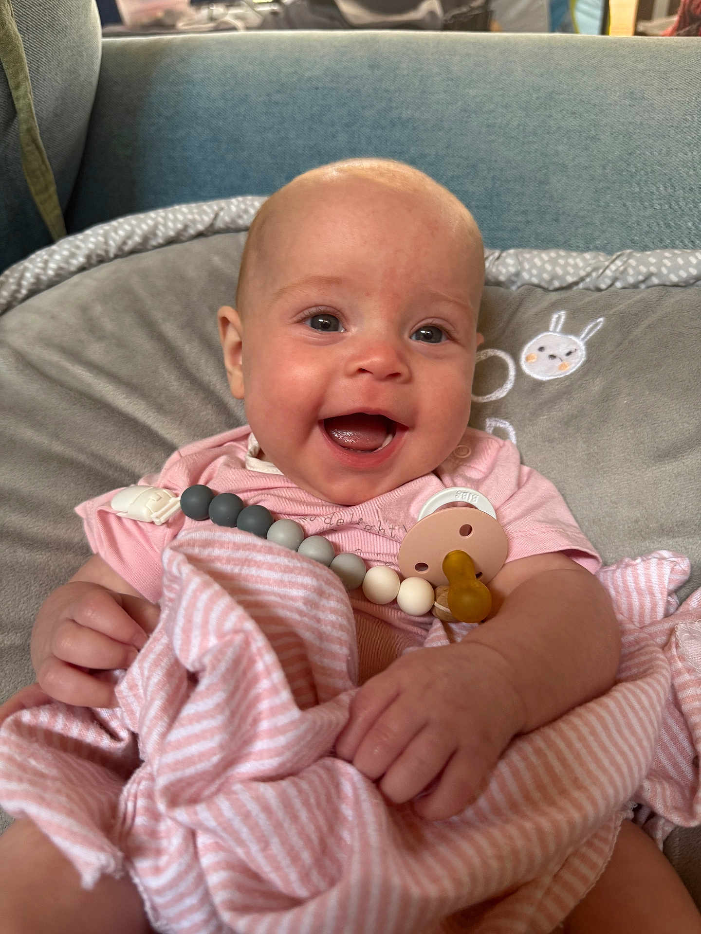 Emmaline joined the competition — help win amazing prizes! baby, smiling, blanket, pacifier, pink_clothing, cushion, gray_cushion, indoor, child, happy, face, hands, seated, infant, cute, soft_toys, comfort, portrait, closeup, home