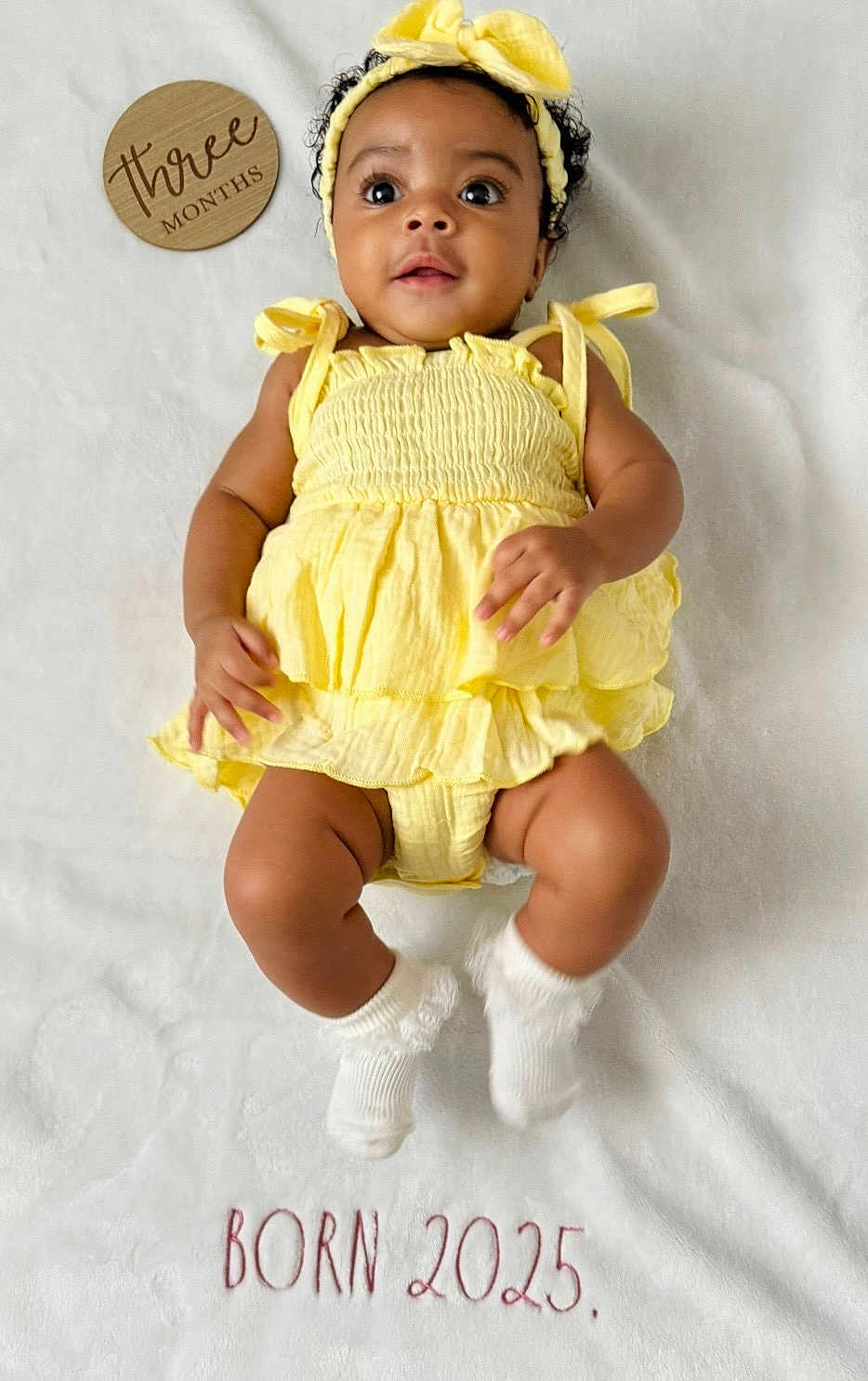 Arya joined the competition — help win amazing prizes! baby, infant, yellow_dress, headband, white_blanket, wooden_plaque, three_months, born_2025, smiling, cute, lying_down, hands, legs, socks, portrait, child, close_up, soft_texture, indoor, happy