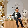 balloons, birthday_cake, boots, cake, celebration, child, decorations, event, festive, gold, happy_birthday, indoor, leather_jacket, party, portrait, smiling, table, toddler, white, young_child