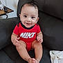 baby, child, smiling, red_clothing, nike, onesie, sitting, leather_chair, indoor, happy, cute, portrait, person, infant, small_table, remote_control, lamp, home, cozy, skin