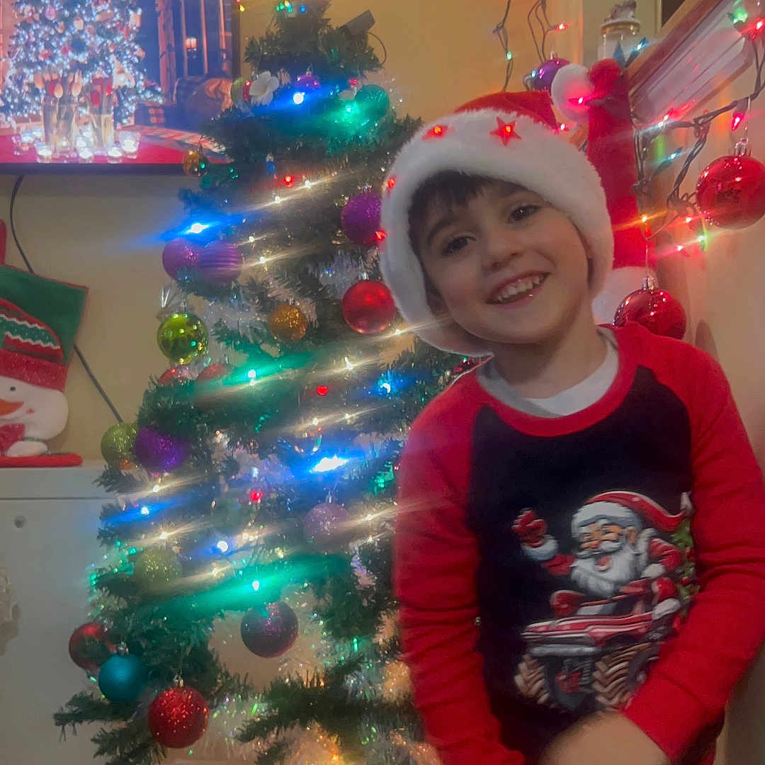 Dany is registered to the contest to win money with this photo: celebration, child, christmas_tree, decorations, festive, green, happy, holiday, home, indoor, joyful, lights, ornaments, pajamas, red, santa_hat, seasonal, smiling, wall, young_child
