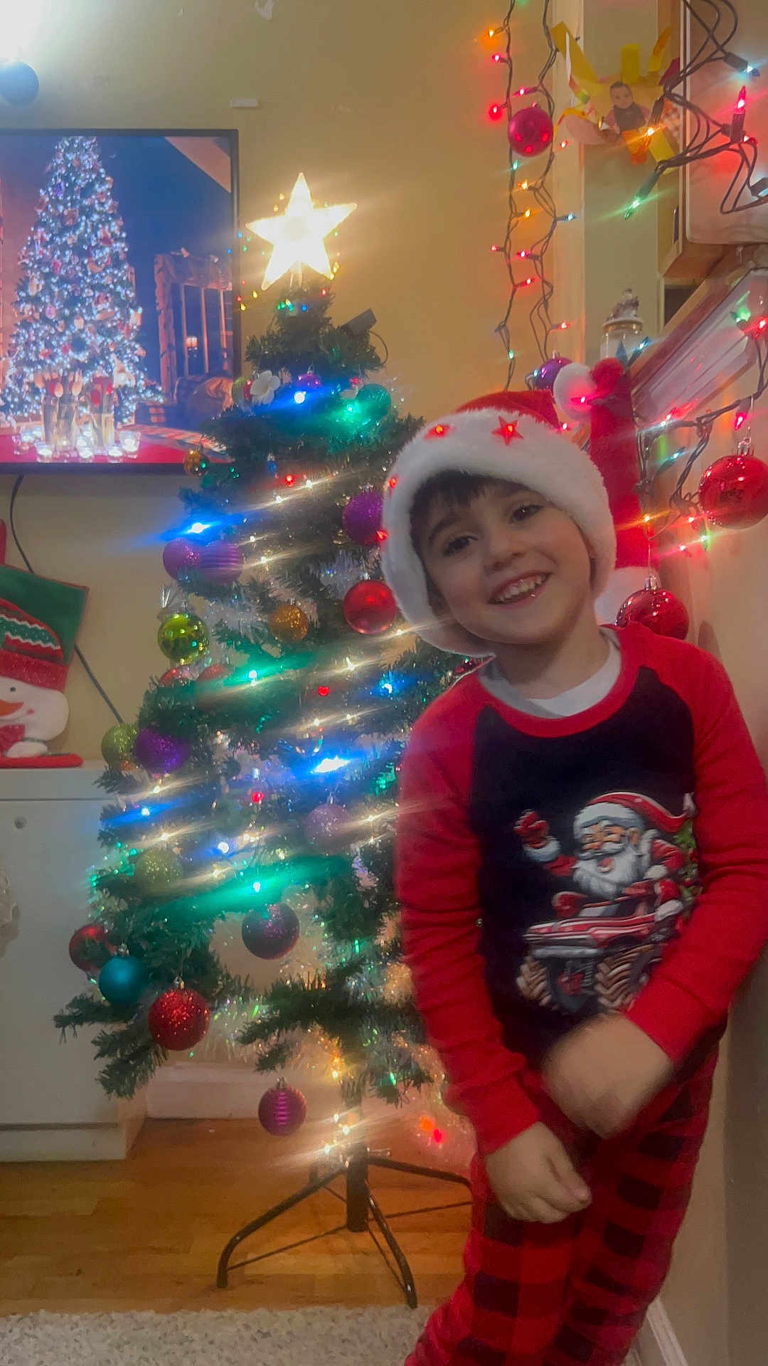 Dany is registered to the contest to win money with this photo: child, christmas_tree, holiday, festive, santa_hat, pajamas, smiling, indoor, decorations, lights, ornaments, red, green, happy, home, celebration, seasonal, young_child, joyful, wall