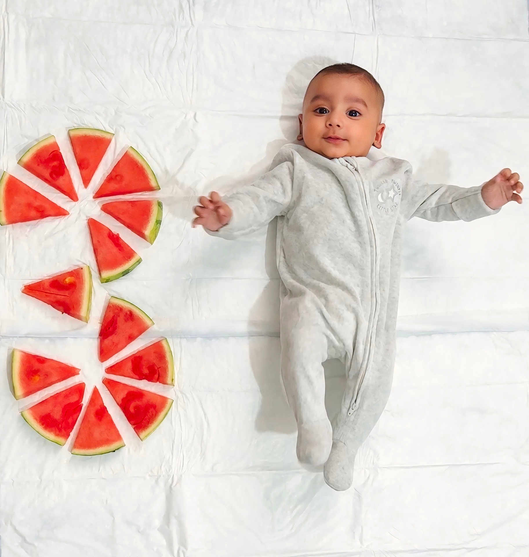Tashfeen is registered to the contest to win money with this photo: baby, infant, watermelon, fruit, slices, white_sheet, onesie, clothing, laying_down, arms_outstretched, cute, fresh, healthy_food, summer, food_arrangement, child, happy, soft_lighting, playful, portrait