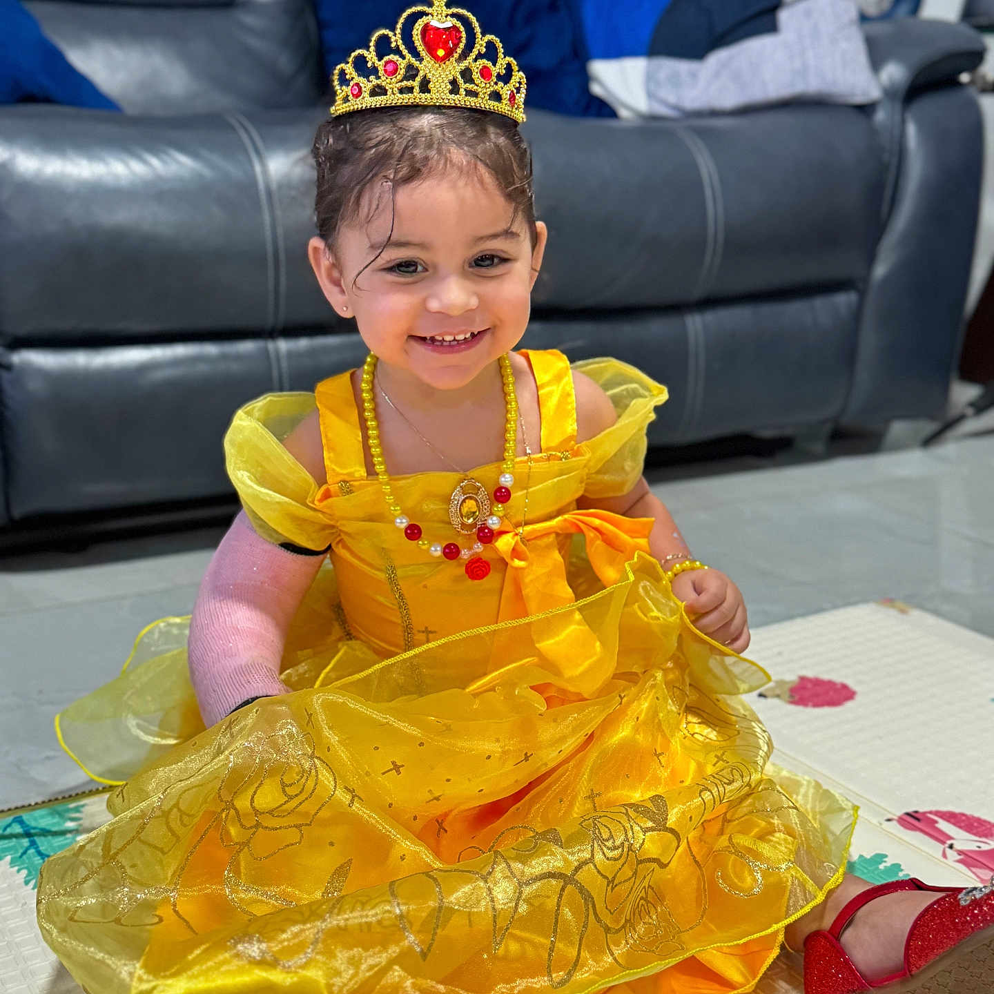Bella is registered to the contest to win money with this photo: arm_cast, cast, child, couch, floor, footwear, girl, happy, indoor, jewelry, leather_couch, necklace, play_mat, portrait, red_shoe, sitting, smiling, tiara, toy, yellow_dress