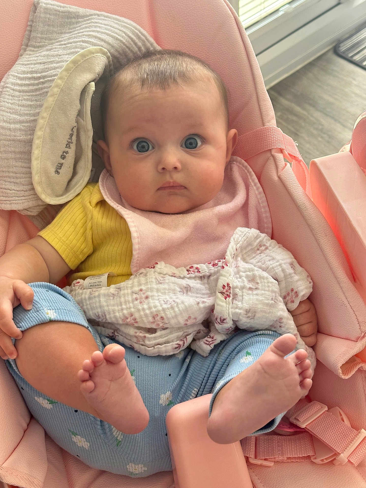 Arabella is registered to the contest to win money with this photo: baby, infant, child, pink_seat, yellow_shirt, blue_pants, bare_feet, blanket, bib, indoors, floor, cushion, person, cute, wide_eyes, soft_toys, comfort, portrait, baby_clothing, holding_leg