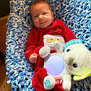Riley joined the competition — help win amazing prizes! baby, infant, red_clothing, santa_design, plush_toy, glowing_toy, knitted_blanket, blue_and_white, smiling, cozy, indoor, wood_floor, cute, festive, holiday, child, soft_toy, portrait, happy, relaxed