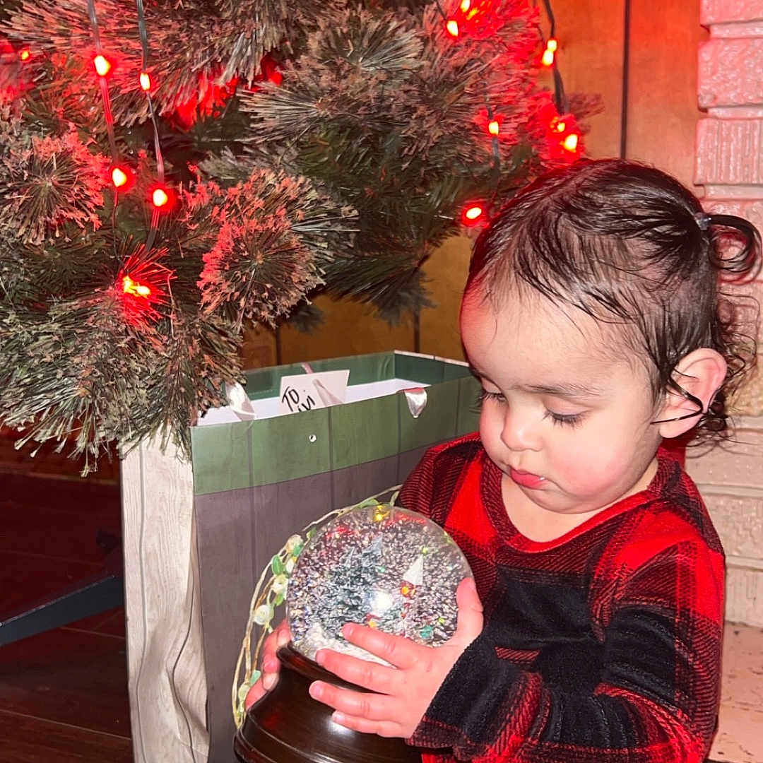 Viviana is registered to the contest to win money with this photo: candlelight, child, christmas_tree, cozy, curious, cute, decorations, face, festive, gift_bag, holiday, holiday_lights, indoors, pajamas, person, red_and_black, snow_globe, toddler, winter, wooden_floor