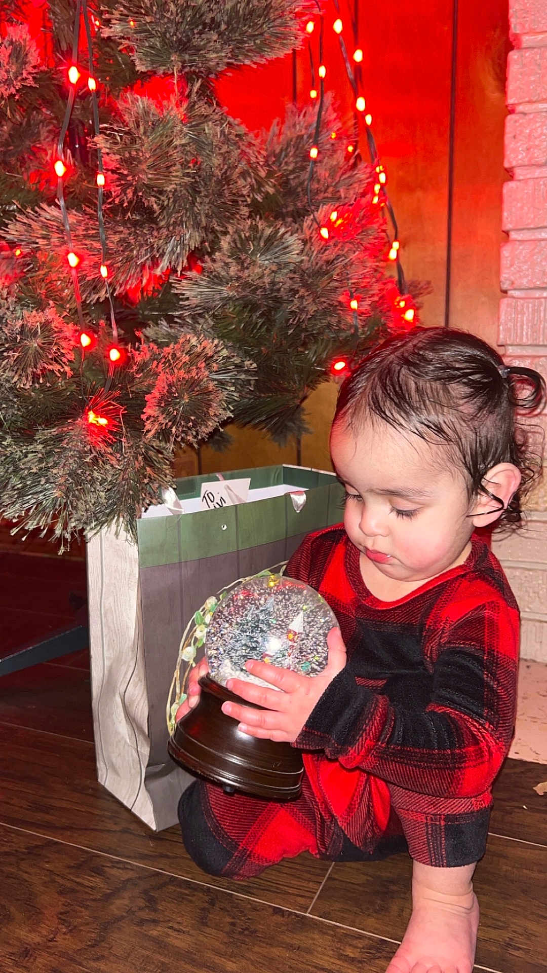 Viviana is registered to the contest to win money with this photo: toddler, child, snow_globe, christmas_tree, holiday_lights, pajamas, red_and_black, gift_bag, curious, indoors, wooden_floor, holiday, decorations, face, person, candlelight, festive, cozy, winter, cute