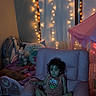 child, pink_chair, curly_hair, string_lights, stuffed_animals, bed, blanket, pillow, tent, playroom, indoor, soft_lighting, cozy, carpet, person, cute, relaxed, decor, toy, nighttime