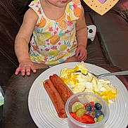 Viviana is registered to the contest to win money with this photo: toddler, child, breakfast, plate, sausages, scrambled_eggs, fruit, grapes, blueberries, strawberries, cup, leather_couch, snack_pouch, dress, food, indoor, person, sitting, heart_sticker, curious