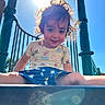 child, toddler, smiling, sunlight, curly_hair, playground, outdoor, blue_sky, green_poles, sunflare, happy, sitting, casual_clothing, shorts, tshirt, daylight, play, fun, young, portrait