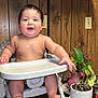 baby, bare_torso, child, cup, drooling, feeding_tray, hat, highchair, hot_dog_buns, hot_sauce_bottle, indoor, plant, plate, potted_plant, shelf, smile, tinsel, toes, wood_paneling, wooden_table