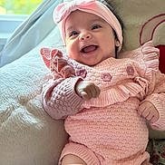 Amreen is registered to the contest to win money with this photo: baby, smiling, pink_clothing, headband, knitted_outfit, cushion, indoor, happy, infant, cute, portrait, soft_texture, cozy, child, person, sitting, face, hand, window, natural_light