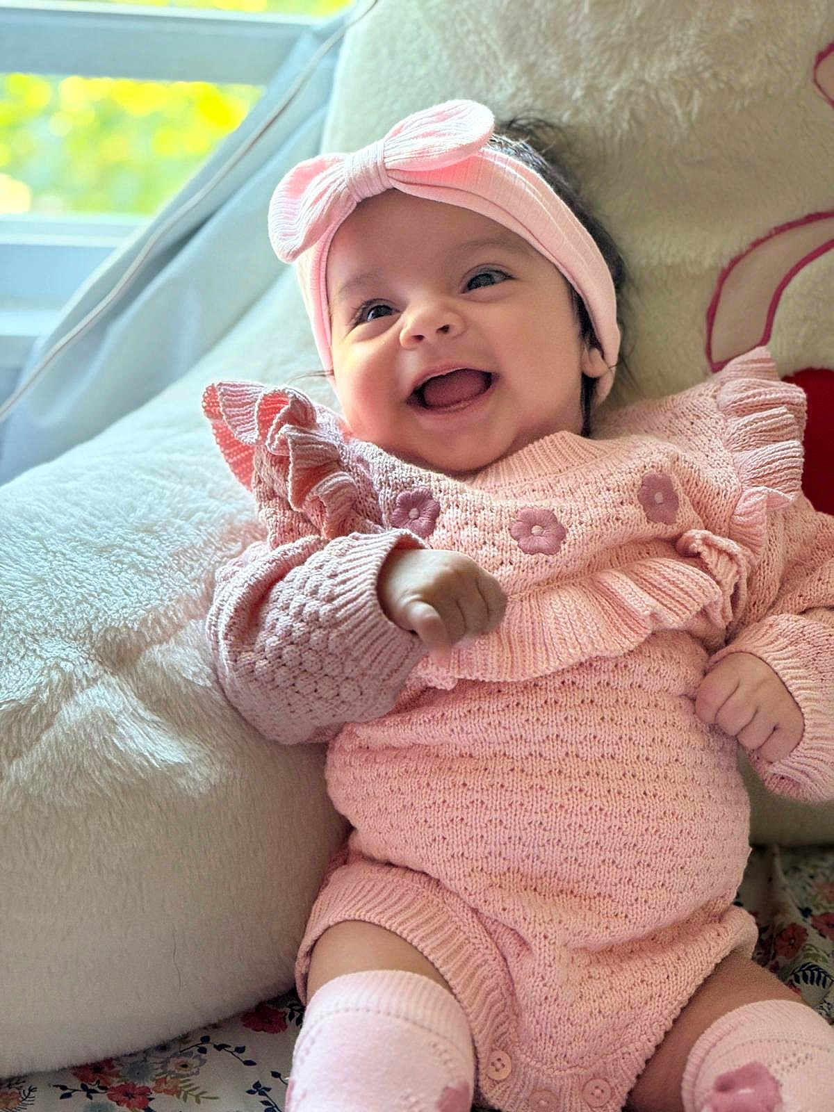 Amreen is registered to the contest to win money with this photo: baby, smiling, pink_clothing, headband, knitted_outfit, cushion, indoor, happy, infant, cute, portrait, soft_texture, cozy, child, person, sitting, face, hand, window, natural_light