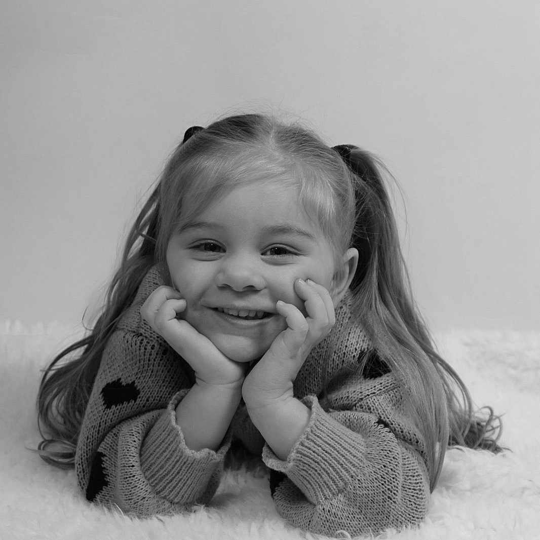 Azëlia participe au concours pour gagner de l'argent avec cette photo : child, girl, smiling, pigtails, sweater, portrait, black_and_white, happy, face, hands, lying_down, blanket, cozy, cute, hair, young, innocent, resting, expression, studio
