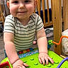 toddler, child, toy, green_toy, smiling, indoor, nursery, crib, baby, playtime, curious, striped_shirt, hands, wooden_furniture, floor, cute, person, infant, play, happy