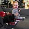 toddler, child, dog, sofa, blanket, pajamas, smiling, indoor, home, living_room, furniture, pet, black_dog, gaming_chair, shelf, person, stairs, cozy, casual, relaxed