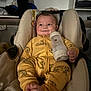 baby, infant, car_seat, bottle, milk, yellow_onesie, tiger_print, smile, feet, hands, face, cheeks, seat_padding, warning_label, clothing, indoor, soft_lighting, portrait, cute, cozy