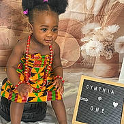 Cynthia joined the competition — help win amazing prizes! toddler, child, smiling, traditional_clothing, beads, hair_bows, shoes, stool, signboard, birthday, portrait, indoor, fabric, decor, person, happy, cute, celebration, flower_background, colorful