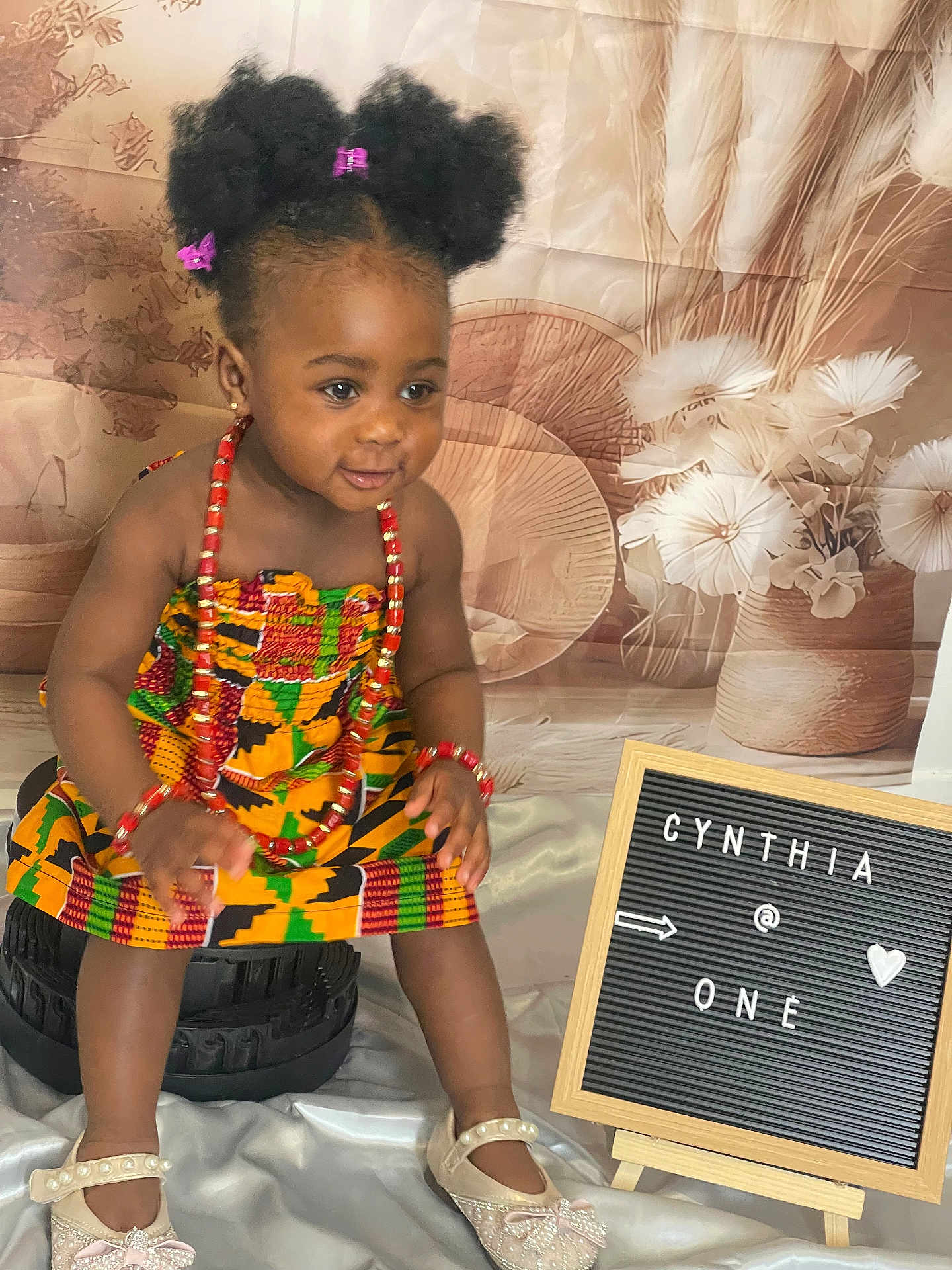 Cynthia joined the competition — help win amazing prizes! toddler, child, smiling, traditional_clothing, beads, hair_bows, shoes, stool, signboard, birthday, portrait, indoor, fabric, decor, person, happy, cute, celebration, flower_background, colorful