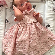 Pebble is registered to the contest to win money with this photo: baby, infant, pink_dress, floral_pattern, lying_down, padded_mat, penguin_print, blanket, cute, child, portrait, playful_expression, hand_near_head, soft_lighting, indoors, white_socks, lace_trim, smiling, face, person