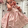 baby, infant, pink_dress, floral_pattern, lying_down, padded_mat, penguin_print, blanket, cute, child, portrait, playful_expression, hand_near_head, soft_lighting, indoors, white_socks, lace_trim, smiling, face, person
