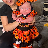 Lottie joined the competition — help win amazing prizes! baby, woman, halloween_costume, orange, black, polka_dots, headband, smile, couch, living_room, person, holding, baby_clothes, footwear, sneakers, floor, blanket, jewelry, happy, indoor