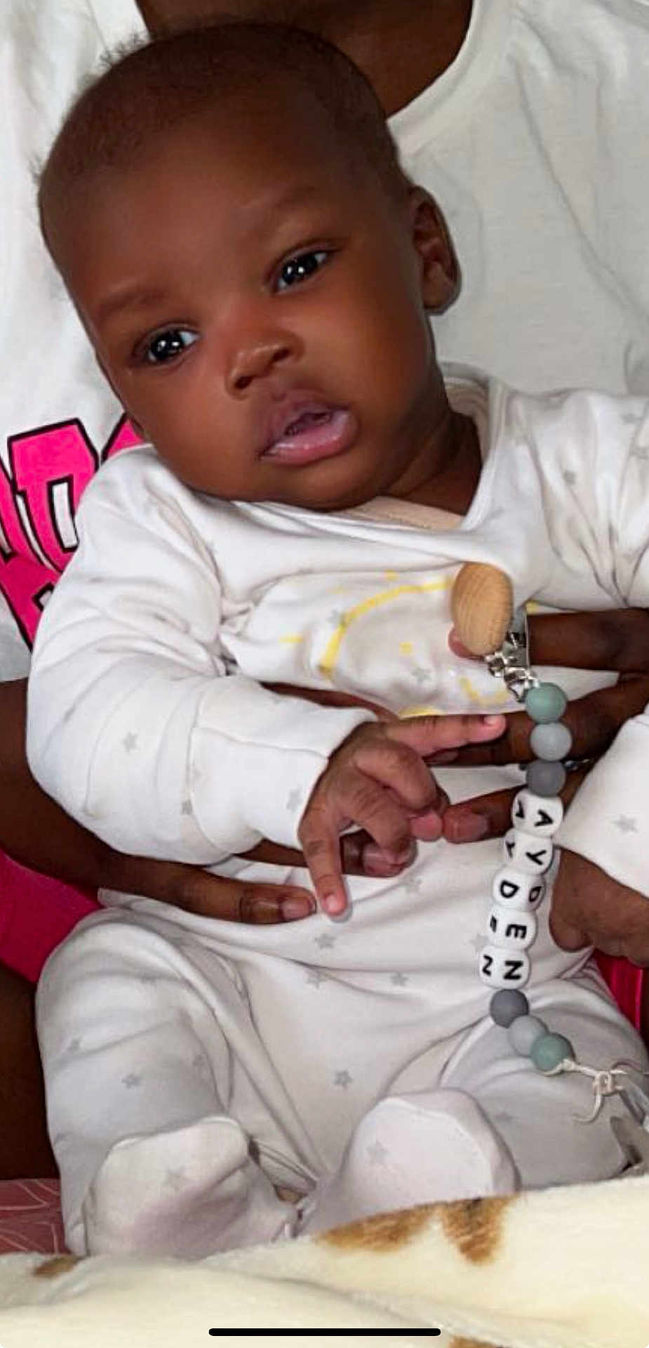 Ayden participe au concours pour gagner de l'argent avec cette photo : baby, infant, child, onesie, pacifier_clip, name_beads, hand, adult_hand, white_clothing, star_pattern, indoor, soft_blanket, person, cute, curious, holding, seated, closeup, face, skin