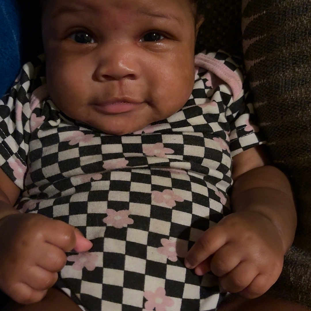 Zari is registered to the contest to win money with this photo: baby, black_and_white, checkered_pattern, child, clothing, couch, cute, expression, face, flower_pattern, hands, indoors, infant, legs, pink, portrait, sitting, skin, texture, young