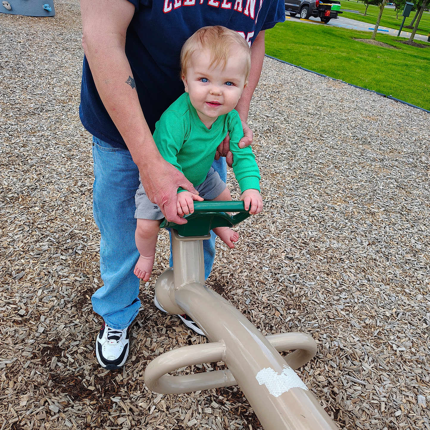 Noah is registered to the contest to win money with this photo: adult, blue_jeans, casual_clothing, child, daylight, fun, grass, green_shirt, holding, outdoor, park, person, play, playground, seesaw, smiling, sneakers, toddler, trees, wood_chips