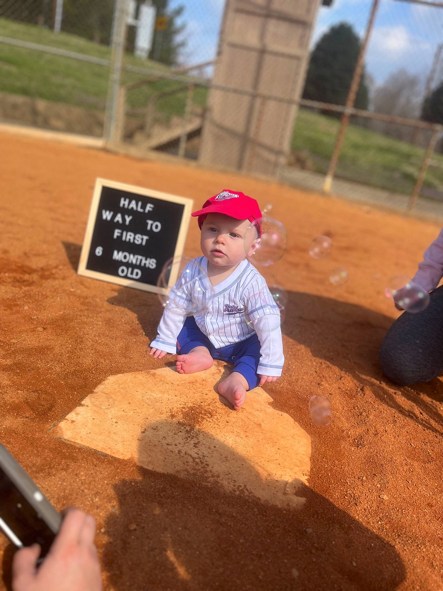 Blake joined the competition — help win amazing prizes! asphalt, baseball_cap, cap, child, flooring, fun, grass, hat, headwear, landscape, leisure, mobile_phone, person, play, recreation, sand, sitting, sky, soil, toddler