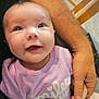 baby, child, smile, purple_shirt, text_on_clothing, adult_arm, hand_holding, chair, floor, tiled_floor, indoor, person, cute, happy, infant, sitting, skin, casual_clothing, portrait, closeup