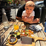 Roman joined the competition — help win amazing prizes! baby, man, outdoor, table, food, drink, plant, glasses, cap, plate, fork, spoon, wood, restaurant, sunlight, baby_held, curious, meal, urban, tattoo