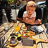 baby, man, outdoor, table, food, drink, plant, glasses, cap, plate, fork, spoon, wood, restaurant, sunlight, baby_held, curious, meal, urban, tattoo