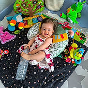 Kiara participe au concours pour gagner de l'argent avec cette photo : baby, child, playmat, toy, pillow, water_bottle, dress, smile, happy, indoor, plush_toy, colorful, cushion, floor_mat, infant, person, cute, play_area, soft_toy, cheerful