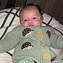 baby, knitted_clothing, green_outfit, hedgehog_pattern, bedspread, indoor, portrait, infant, cute, toddler, buttoned_sweater, lying_down, face, person, home, warm_clothing, soft_texture, hair, expression, resting