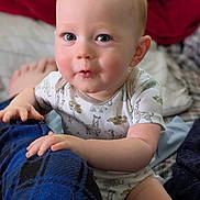 Hudson is registered to the contest to win money with this photo: baby, child, infant, onesie, animal_print, blanket, blue_blanket, hand, face, eyes, skin, red_background, bed, comfort, cute, expression, indoor, person, portrait, soft_lighting