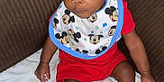 Andreus is registered to the contest to win money with this photo: baby, child, mickey_mouse, bib, red_clothing, socks, couch, blanket, indoor, curly_hair, person, infant, seated, looking_away, cute, young_child, expression, casual_clothing, home, soft_fabric