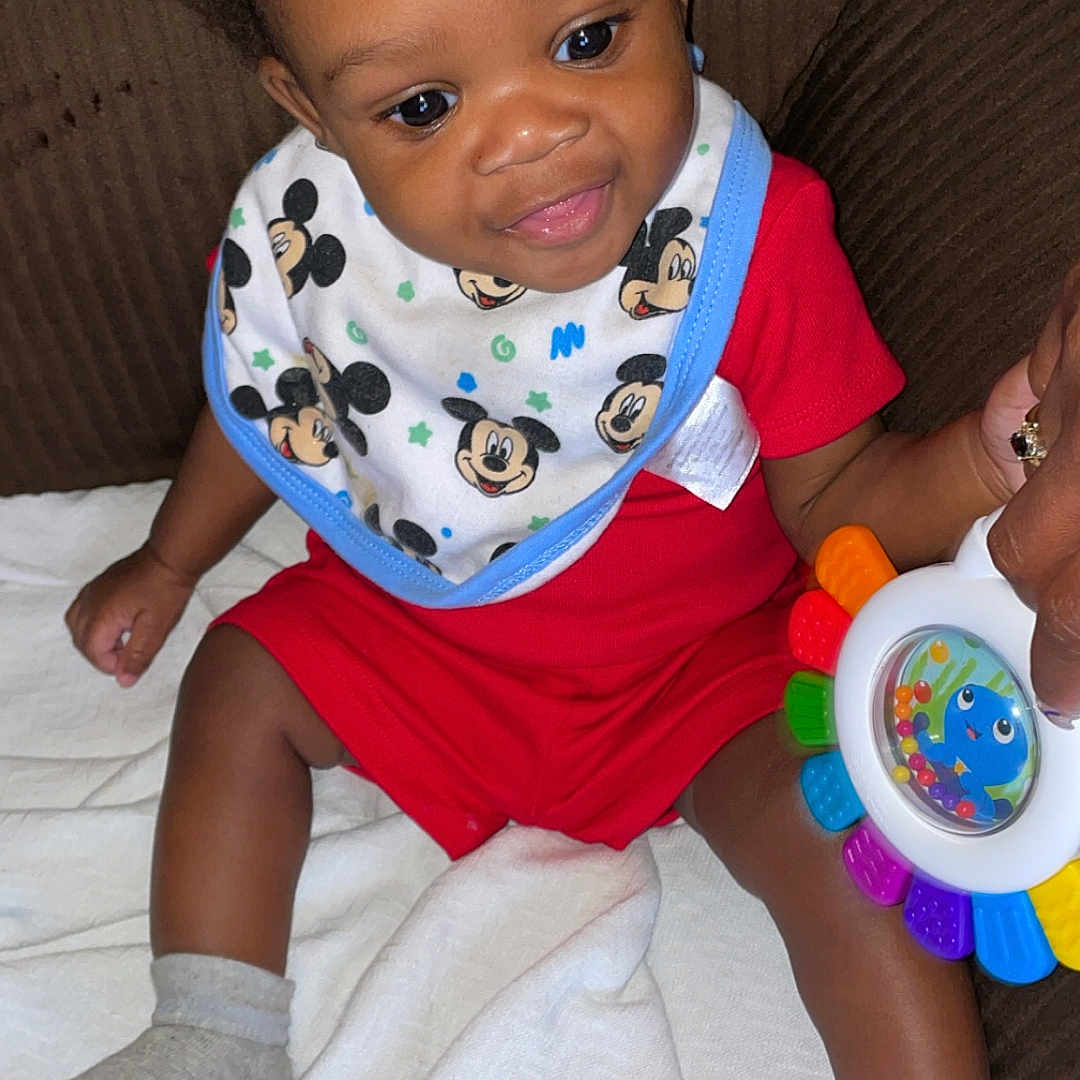 Andreus is registered to the contest to win money with this photo: baby, bib, blanket, child, colorful, couch, cute, face, hand, happy, indoor, infant, mickey_mouse, person, playful, red_clothing, sitting, smiling, sock, toy